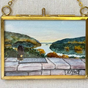 May include: A rectangular watercolor painting in a gold metal frame with a hanging chain. The artwork depicts a person with dark hair sitting on a stone wall overlooking a calm lake surrounded by green hills and trees. The signature RPat is visible.