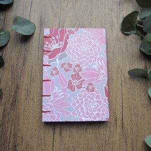 Pode incluir: Um caderno com padrão floral em tons de rosa e cinza. A capa apresenta um design repetido de flores estilizadas em tons de rosa e branco. A lombada é encadernada com fio vermelho.