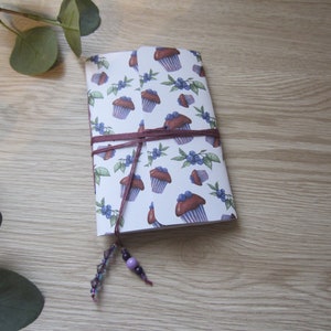 Peut inclure: Un carnet blanc avec un motif de cupcakes et de myrtilles violets et bruns. Le carnet est attaché avec une ficelle brune et a un pompon à perles violet et argent.