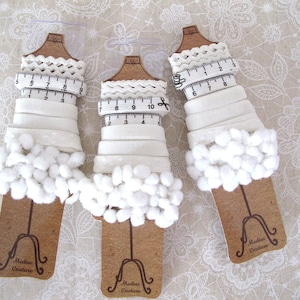 May include: Three spools of white trim with white pom poms on the bottom. Each spool has a white measuring tape wrapped around it. The spools are labelled "Medias Couture".
