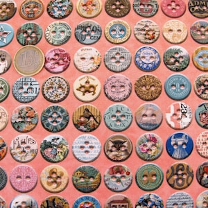 Op de afbeelding: Een verzameling kleurrijke, ronde knopen met verschillende ontwerpen en patronen. Sommige knopen bevatten tekst, waaronder "Broken", "25 Cents", "Flowers", "Shoe Store" en "Good For 10 Cents".