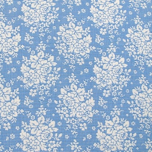 Könnte beinhalten: Nahaufnahme eines blauen Stoffes mit weißem Blumenmuster. Das Design zeigt Ansammlungen weißer Rosen und kleinerer Blüten, vor einem hellblauen Hintergrund mit einer subtilen gepunkteten Textur. Der Stoff hat eine Vintage- oder Cottagecore-Ästhetik.