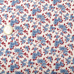 May include: A white fabric featuring a repeating pattern of small blue flowers and dark red leaves on thin brown stems. A gold-coloured one-euro coin is placed on the fabric to provide a sense of scale for the floral print.