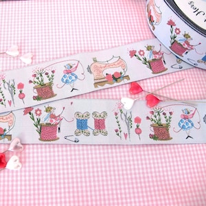 Op de afbeelding: Lichtblauw lint met een herhalend patroon van illustraties met naaithema's. Het ontwerp omvat feeën, garenklosjes, naaimachines en bloemen. Het lint wordt gepresenteerd op een roze ruitjesachtergrond met hartvormige spelden.