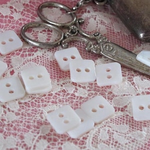Può includere: Una collezione di bottoni quadrati bianchi con due fori ciascuno, sparsi su una superficie di pizzo rosa. Sono visibili anche forbici d'argento e un piccolo oggetto metallico, che suggeriscono un tema di cucito o artigianato.