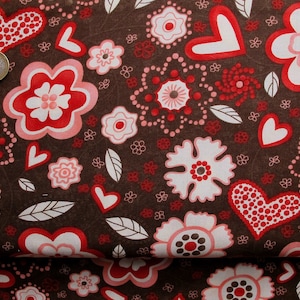 Op de afbeelding: Bruine stof met een herhalend patroon van roze en rode bloemen, harten en bladeren. De harten zijn gevuld met rode stippen.