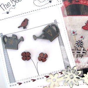 May include: Wooden craft embellishments including a bird, watering cans, and flowers. The bird is brown with red polka dots. The watering cans are grey with gold accents. The flowers are red with brown polka dots.