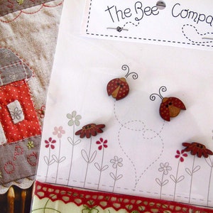Può includere: Un biglietto bianco con il testo "The Bee Company" stampato su di esso. Il biglietto è decorato con bottoni a forma di coccinella rossi e marroni, e un cuore disegnato a puntini. Il biglietto è circondato da un bordo di pizzo rosso e bianco.