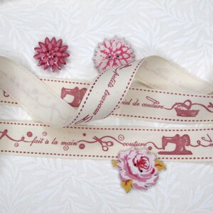 Può includere: Un nastro in tessuto color crema con una stampa rossa ripetuta di macchine da cucire, forbici, rocchetti e il testo "fait à la main" e "matériel de couture". Tre bottoni a forma di fiore nei toni del rosa e del rosso sono posti sul nastro.