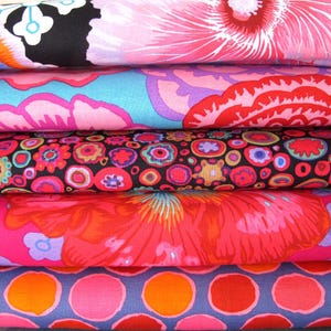 Op de afbeelding: Een stapel kleurrijke stoffen met bloemen- en stippenpatronen. De stoffen hebben verschillende kleuren, waaronder roze, oranje, blauw en zwart. De bloempatronen omvatten grote, gestileerde bloemen en kleinere, meer ingewikkelde ontwerpen.
