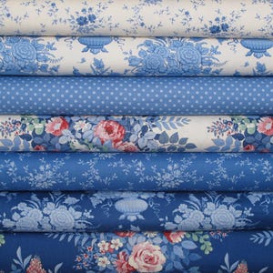 Könnte beinhalten: Ein Stapel aus sieben gefalteten Stoffballen mit verschiedenen blau-weißen Mustern. Die Designs umfassen Blumenarrangements, Urnen, kleine Punkte und zarte Rankenmotive. Die Farbpalette besteht aus verschiedenen Blau-, Weiß- und Rosatönen.