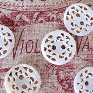 Op de afbeelding: Zes witte, ronde knopen met ingewikkelde, opengewerkte ontwerpen. Elke knoop heeft twee gaten en een bloemachtig patroon dat in het oppervlak is gesneden. De knopen zijn gerangschikt op een textiel met het woord "WATER" zichtbaar.