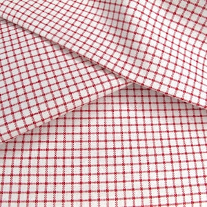 Könnte beinhalten: Nahaufnahme eines weißen Stoffes mit rotem Karomuster. Der Stoff ist gefaltet und zeigt seine Textur und sein Design. Das Muster besteht aus kleinen, gleichmäßig verteilten Quadraten, die einen klassischen Look erzeugen. Ideal für Heimdekoration oder Bastelprojekte.