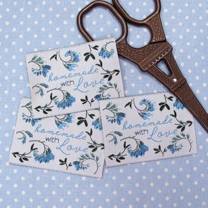 Può includere: Tre etichette in tessuto bianco con motivi floreali blu e il testo "homemade with love" in scrittura corsiva blu. Le etichette sono su uno sfondo blu chiaro con pois bianchi.
