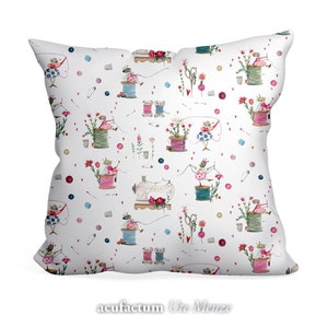 May include: White square pillow with a repeating pattern of sewing-themed illustrations. The design includes spools of thread, sewing machines, scissors, and whimsical figures. Accents of pink, blue, and green are scattered across the pillow's surface. The text "acufactum Ute Menze" is at the bottom.