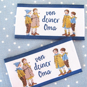 Könnte beinhalten: Zwei weiße Stoff-Etiketten mit blauem Rand, jeweils mit einer bunten Illustration von zwei Kindern und dem Text "von deiner Oma".