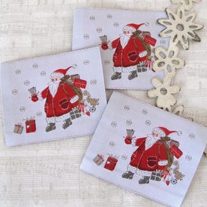 Könnte beinhalten: Drei weiße Stoff-Geschenk-Etiketten mit einer roten und weißen Weihnachtsmann-Illustration, die eine Glocke und einen Sack mit Geschenken hält. Der Hintergrund ist ein weißer Stoff mit einem Schneeflockenmuster.