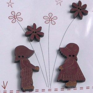 Könnte beinhalten: Holzknöpfe in Form von Kindern und Blumen. Die Knöpfe sind dunkelrot mit einem Used-Look-Finish. Die Kinder tragen Hüte und Kleider. Die Blumen haben sechs Blütenblätter. Der Hintergrund ist weiß mit roten Strichzeichnungen.