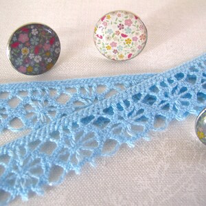 Puede incluir: Primer plano de un encaje de crochet azul con tres cabujones redondos con motivos florales. Los cabujones son plateados y tienen una variedad de flores de colores.