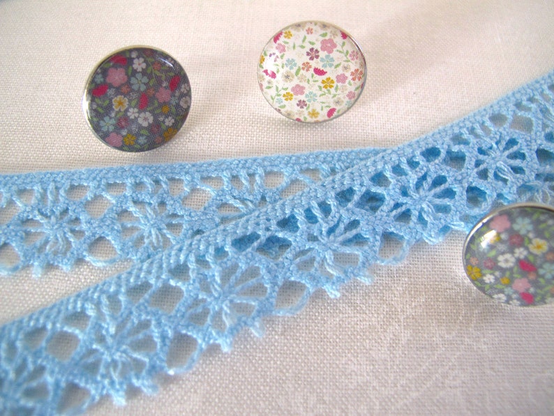Puede incluir: Primer plano de un encaje de crochet azul con tres cabujones redondos con motivos florales. Los cabujones son plateados y tienen una variedad de flores de colores.