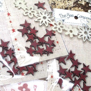 May include: Craft embellishments in clear packaging, including red stars and white flower shapes. The packages are labelled "The Bee Company". The stars are approximately 2.5 cm in size, and the flowers are about 4 cm across. The background is a burlap-like material.