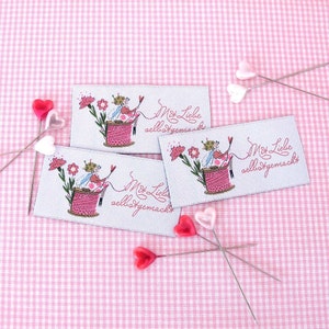 Könnte beinhalten: Drei weiße Stoff-Etiketten mit rosa und grünen Illustrationen einer Fee, die auf einer Garnrolle sitzt. Jedes Etikett trägt den Text "Mit Liebe selbstgemacht" in schwarzer Kursivschrift.