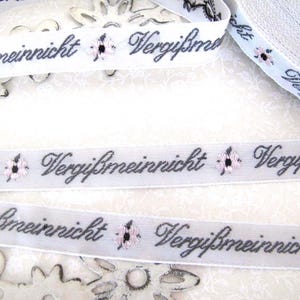 May include: White ribbon with the text "Vergiss mein nicht" in black script and pink flowers embroidered on it.