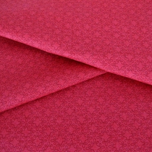 Könnte beinhalten: Nahaufnahme eines roten Stoffes mit einem zarten, sich wiederholenden Muster aus kleinen, wirbelnden Designs. Der Stoff hat eine glatte, weiche Textur.