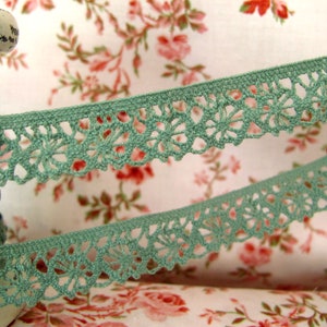 Könnte beinhalten: Eine Spule mit Salbeigrüner Häkelspitze mit einem zarten Blumenmuster. Die Spitze wird vor einem floralen Stoffhintergrund gezeigt.