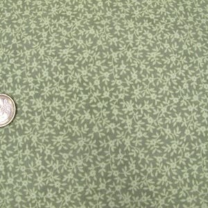 Op de afbeelding: Close-up van een stof met een herhalend bloemenpatroon in lichtgroen en gebroken wit. Een munt van één euro is zichtbaar, als maatreferentie. De stof heeft een delicate, vintage uitstraling, geschikt voor quilt- of knutselprojecten.