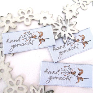 Könnte beinhalten: Drei weiße Stoff-Etiketten mit braunen Blumenmustern und dem Text "hand gemacht" in silberner Schrift.