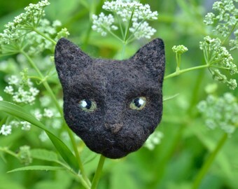 May include: A black felt cat head with white eyes, a black nose, and a fuzzy texture. The cat head is sitting on a bed of green leaves and white flowers.