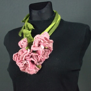 Puede incluir: Un collar de flores de fieltro rosa con hojas de fieltro verde. El collar está en un maniquí negro.