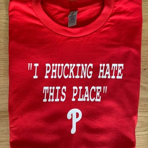 May include: Red t-shirt with white text that reads "I PHUCKING HATE THIS PLACE" and a white Philadelphia Phillies logo.
