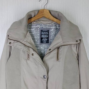 Peut inclure: Une veste beige avec une fermeture &eacute;clair et des boutons. La veste a un col en velours c&ocirc;tel&eacute; et une doublure &agrave; imprim&eacute; journal. L'&eacute;tiquette de la marque indique "Von Dutch" et "True Dutch".