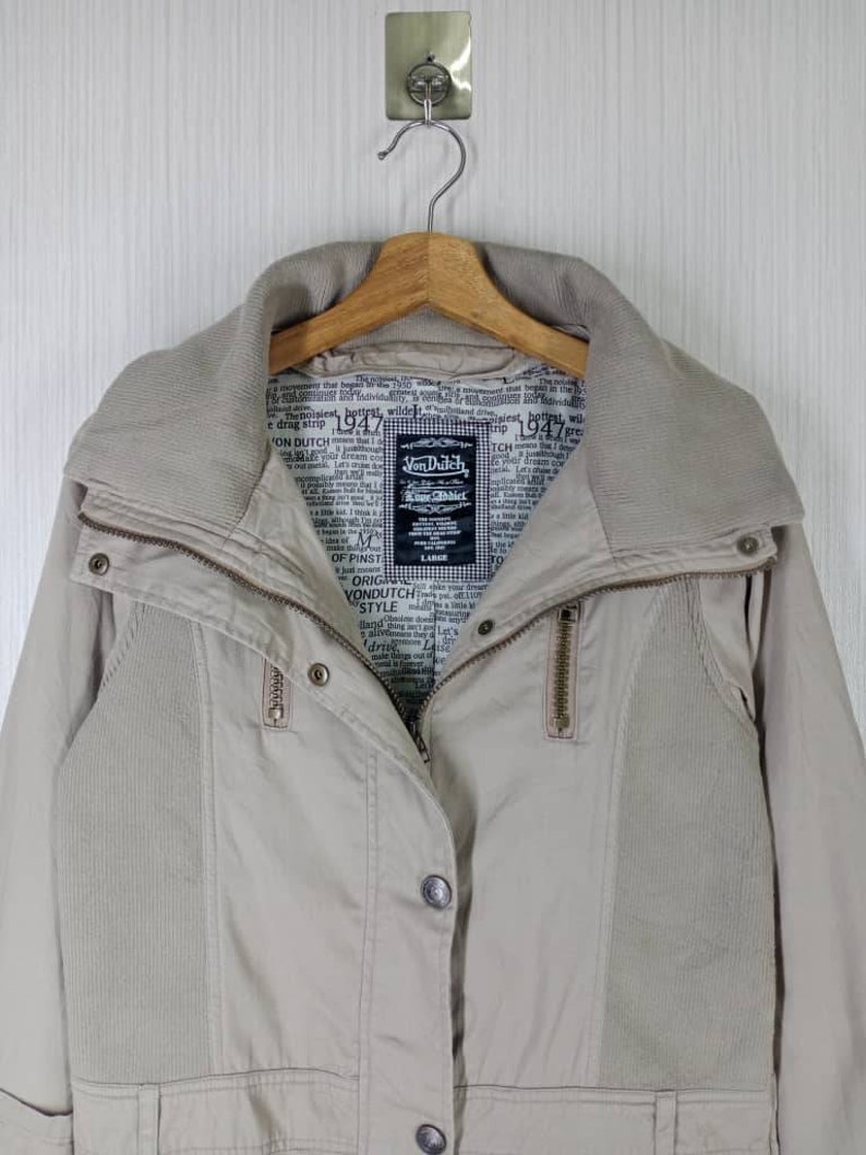 Peut inclure: Une veste beige avec une fermeture &eacute;clair et des boutons. La veste a un col en velours c&ocirc;tel&eacute; et une doublure &agrave; imprim&eacute; journal. L'&eacute;tiquette de la marque indique "Von Dutch" et "True Dutch".