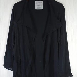 Op de afbeelding: Een zwarte jas met lange mouwen en een gedrapeerde kraag, hangend aan een houten hanger. De jas heeft een vloeiend, lichtgewicht ontwerp. Een klein wit label met zwarte tekst is zichtbaar bij de kraag.