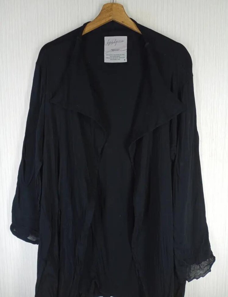 Op de afbeelding: Een zwarte jas met lange mouwen en een gedrapeerde kraag, hangend aan een houten hanger. De jas heeft een vloeiend, lichtgewicht ontwerp. Een klein wit label met zwarte tekst is zichtbaar bij de kraag.