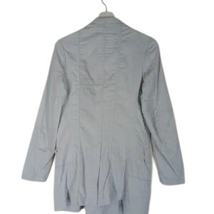 May include: A light gray blazer with long sleeves, hanging from a metal hanger. The blazer has a tailored fit with seams and a slightly asymmetrical hemline. The fabric appears to be a lightweight material.