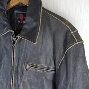 May include: A vintage, distressed leather jacket in a dark grey-blue hue. Features a zip-up front, a collared neckline, and a small chest pocket. The jacket shows signs of wear, with lighter color accents along the seams and edges.