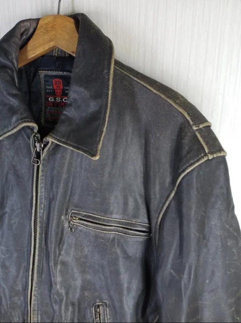 May include: A vintage, distressed leather jacket in a dark grey-blue hue. Features a zip-up front, a collared neckline, and a small chest pocket. The jacket shows signs of wear, with lighter color accents along the seams and edges.