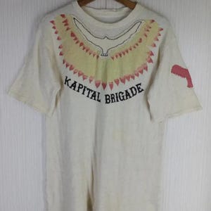 Puede incluir: Camiseta de color crema con un diseño de pájaro y el texto "KAPITAL BRIGADE" en negro. La camiseta tiene cuello redondo y mangas cortas. Un diseño rojo está en la manga derecha.