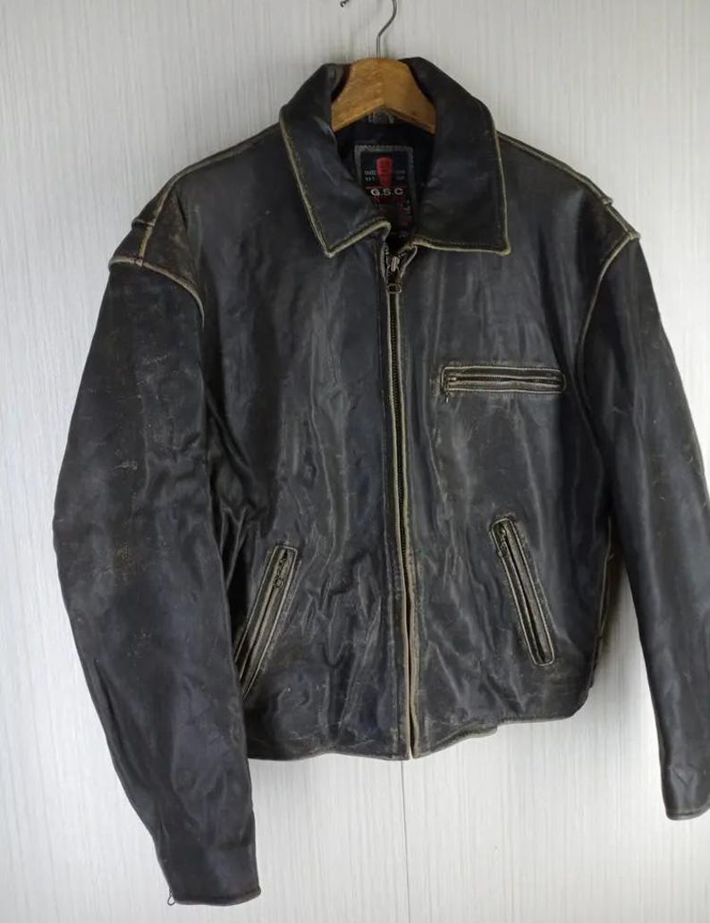 May include: A distressed black leather jacket with a zip-up front, a collared neckline, and multiple pockets. The jacket has a worn, vintage appearance with visible wear and tear, and is hanging from a wooden hanger.