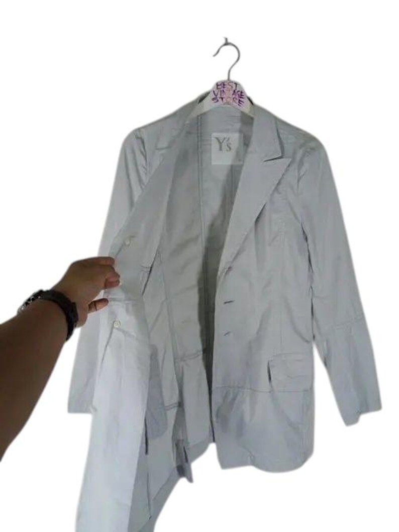 May include: A light gray blazer with a two-button closure, two front pockets, and a notched lapel. The jacket is hanging on a hanger with the text "BEST VINTAGE STORE" and the brand "Y's".