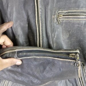 May include: Close-up of a distressed, dark gray leather jacket. Features include a zippered front, a zippered sleeve cuff, and a small zippered chest pocket. The jacket shows signs of wear and tear.