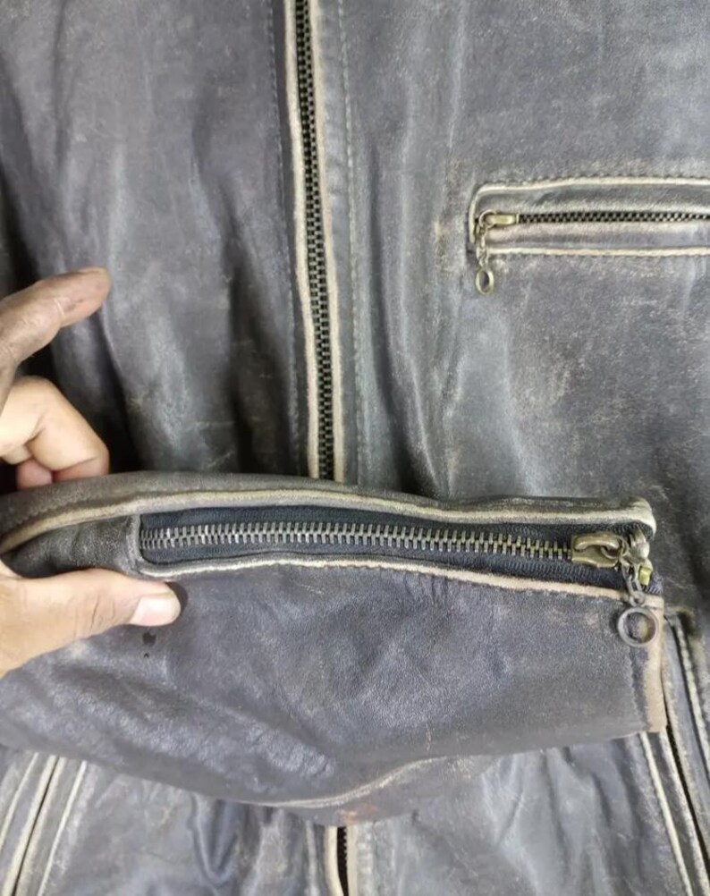 May include: Close-up of a distressed, dark gray leather jacket. Features include a zippered front, a zippered sleeve cuff, and a small zippered chest pocket. The jacket shows signs of wear and tear.