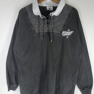May include: A long-sleeve charcoal gray rugby shirt featuring a white collar, a quilted chest panel, and a lace-up placket. The shirt displays an embroidered white fern leaf graphic with the letters N and Z positioned above and below the leaf.