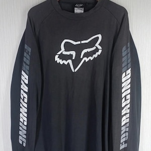 May include: A charcoal gray long-sleeve shirt featuring a large white fox head logo on the chest. The text FOX RACING INC is printed in white down both sleeves, and a small rectangular patch with the text FOX RACING INC is on the lower front.