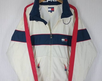 Op de afbeelding: Crèmekleurige jas met marineblauwe en rode accenten. De jas heeft een volledige ritssluiting, een hoge kraag en het Tommy Hilfiger-logo. Het ontwerp omvat rode strepen langs de mouwen en een marineblauwe band over de borst.