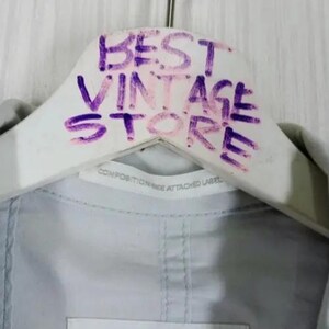 May include: A white wooden hanger with the words "BEST VINTAGE STORE" written in purple marker. The hanger is holding a light gray garment. A label with text is visible.